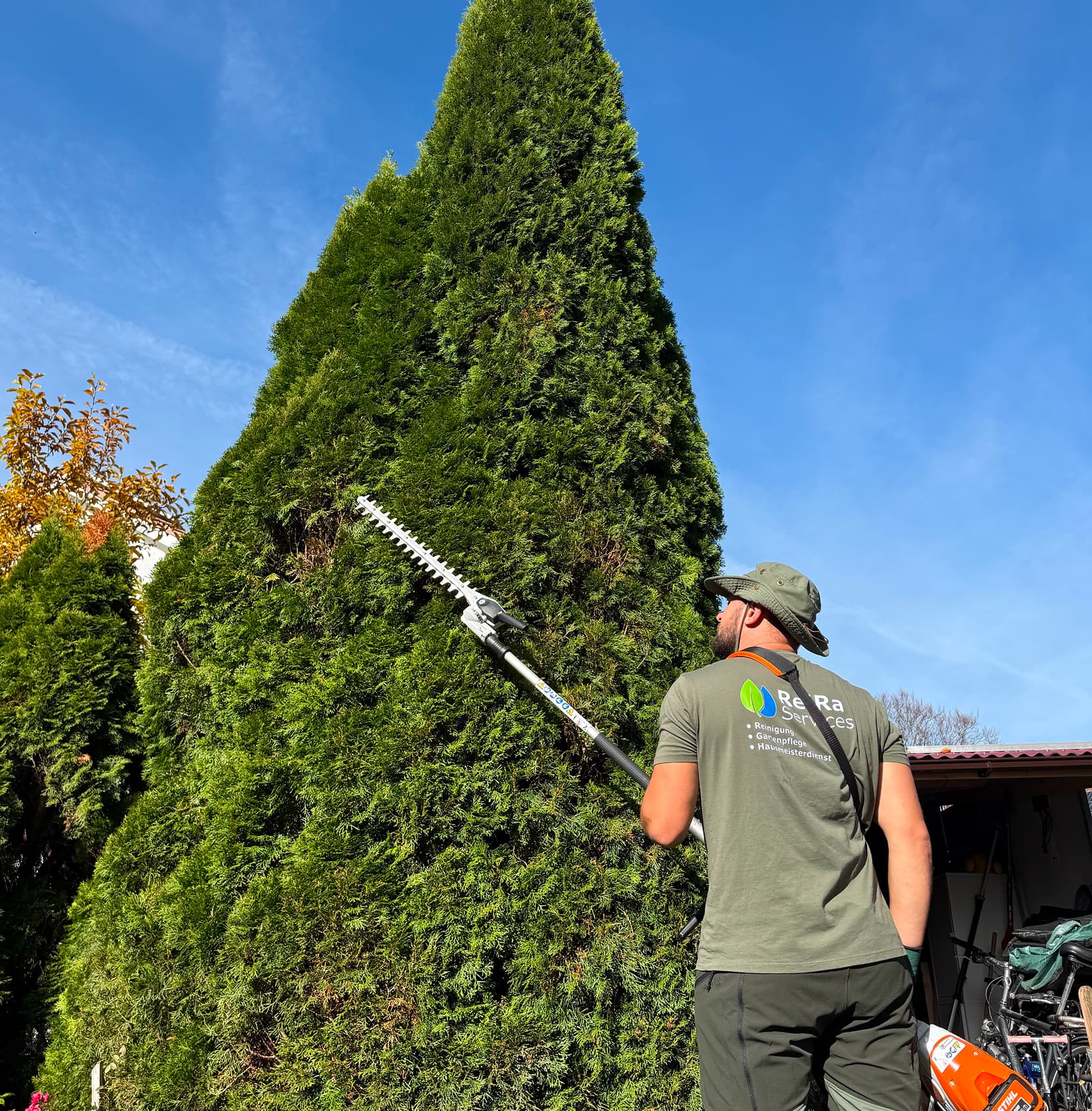Mitarbeiter von ReiRa-Services pflegt eine hohe Hecke mit einem professionellen Heckenschneider unter blauem Himmel.