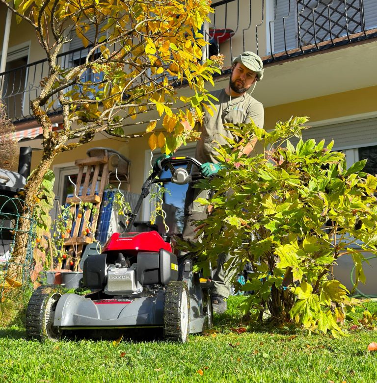 Mitarbeiter von ReiRa-Services mäht den Rasen mit einem Rasenmäher neben einem Wohnhaus und kleinen Bäumen.